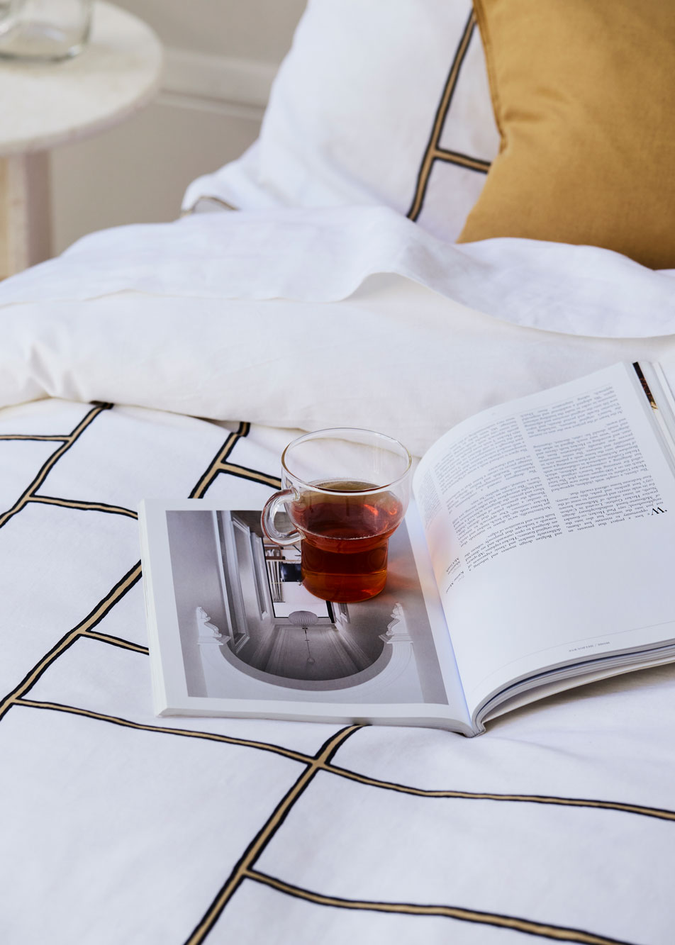 A close-up of a bed. On top of the bed sits an open book with a cup of tea balanced on the open page.