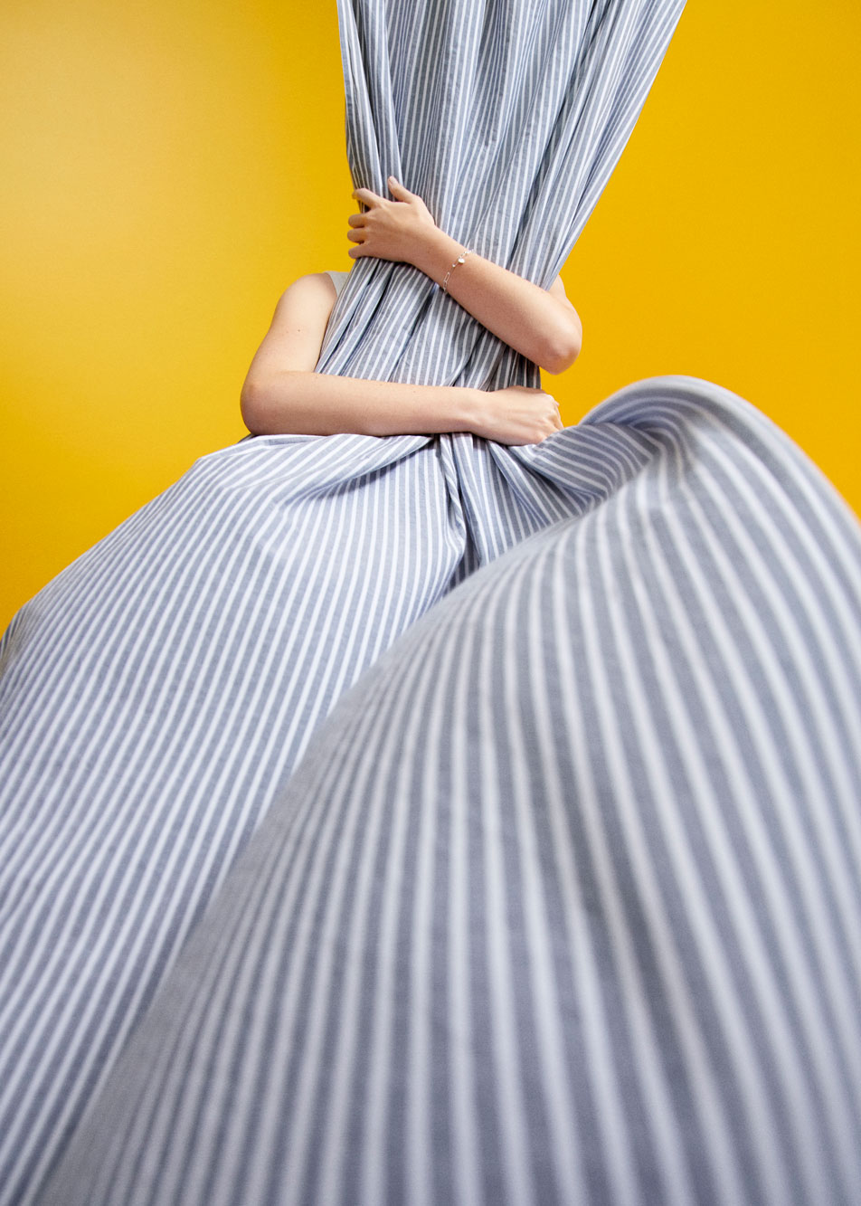 A striped sheet flowing from the ceiling to the floor. A person stands hidden behind it with only their arms showing, wrapped around the sheet. The background is a bright yellow wall.