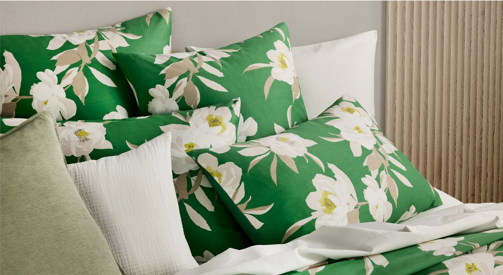 landscape photo of a styled bed, at the head. A glimpse of the quilt and flat sheet is seen, but it is mainly stacked pillows dressed in a lively green with cream oversized peony pattern. two white pillows can be seen peeking throughout.