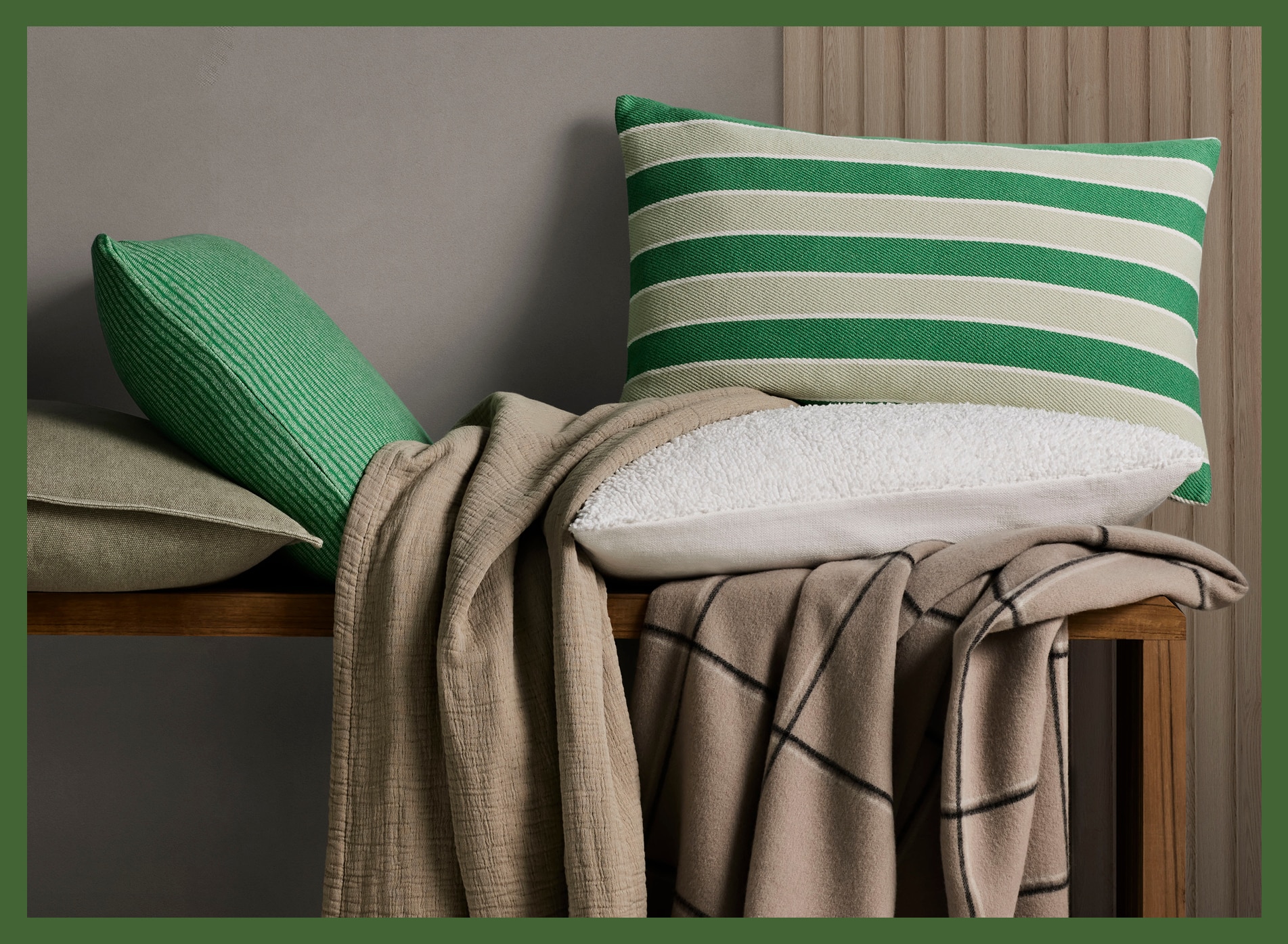 green home decor in styled shot. four pillows, two green stripes, one khaki one white are styled on top of two throws, one a textured brown, one a khaki check with black and white lines. products sit on a wooden bench. image is surrounded by green border.