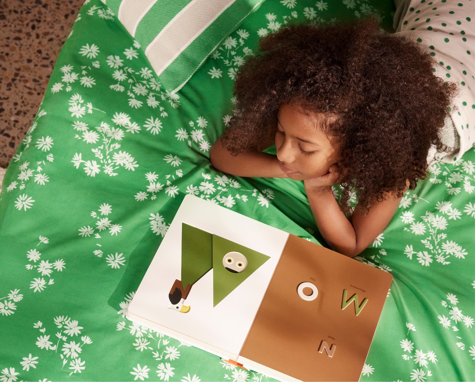 young brown skinned girl with tight, brown curly hair lies on her stomach, propped up on elbows. she is looking at a picture book open to mountains. the bed has green sheets with a white daisy print scattered across it.