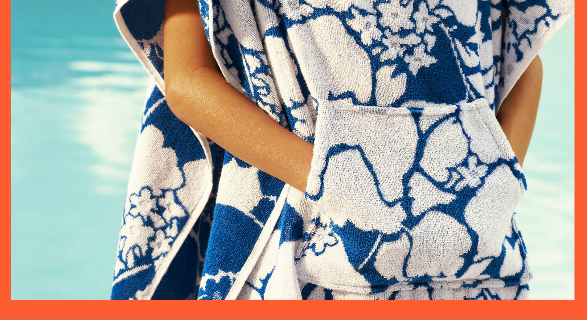 close up of child standing in pool, wearing a white and blue poncho, with hands in pocket. an orange border surrounds image on three sides.
