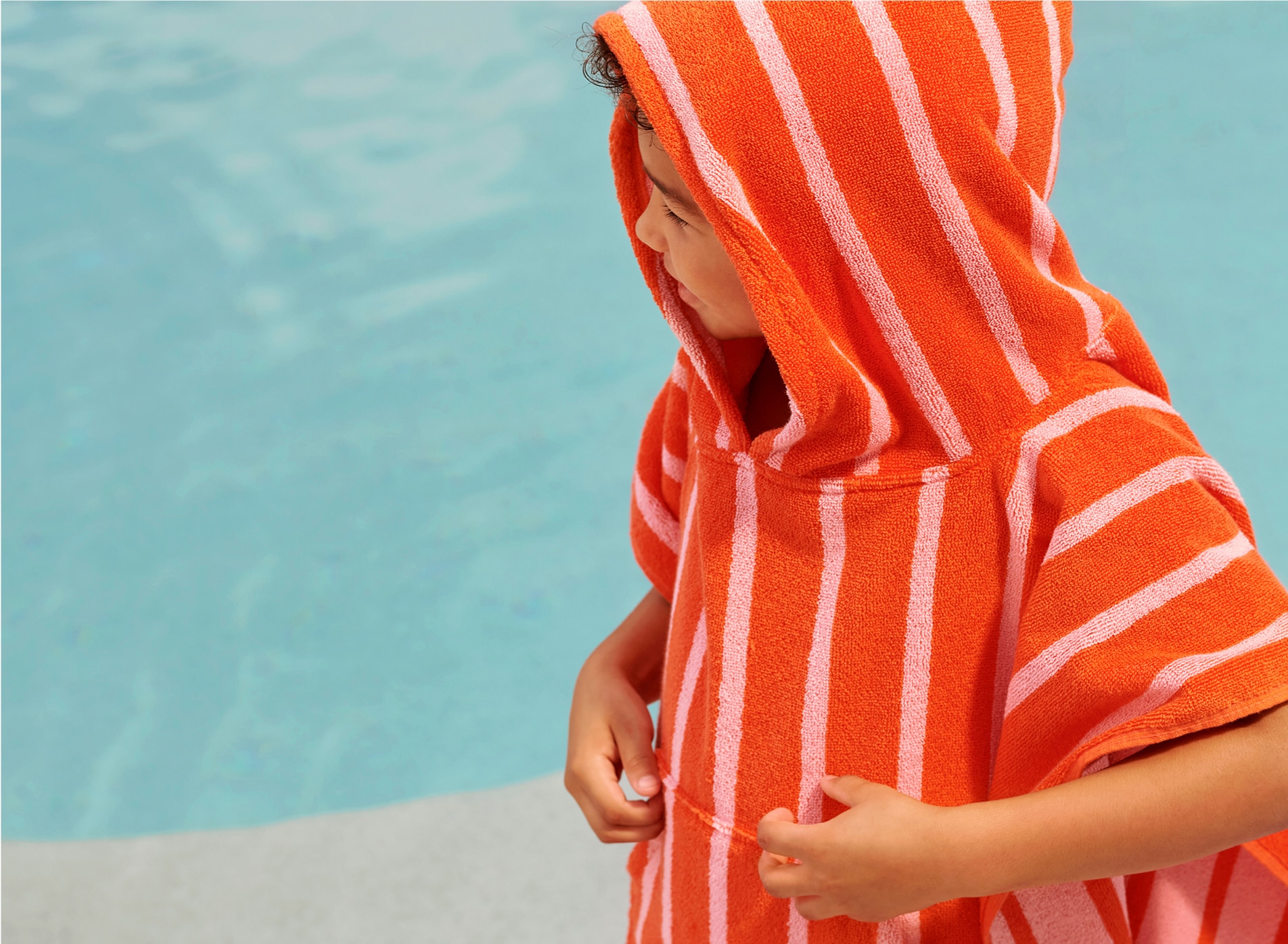 little brown boy with brown curly hair stands on step of pool, looking right. he wears a orange-red poncho with pink stripes, and is touching the fabric on the front.