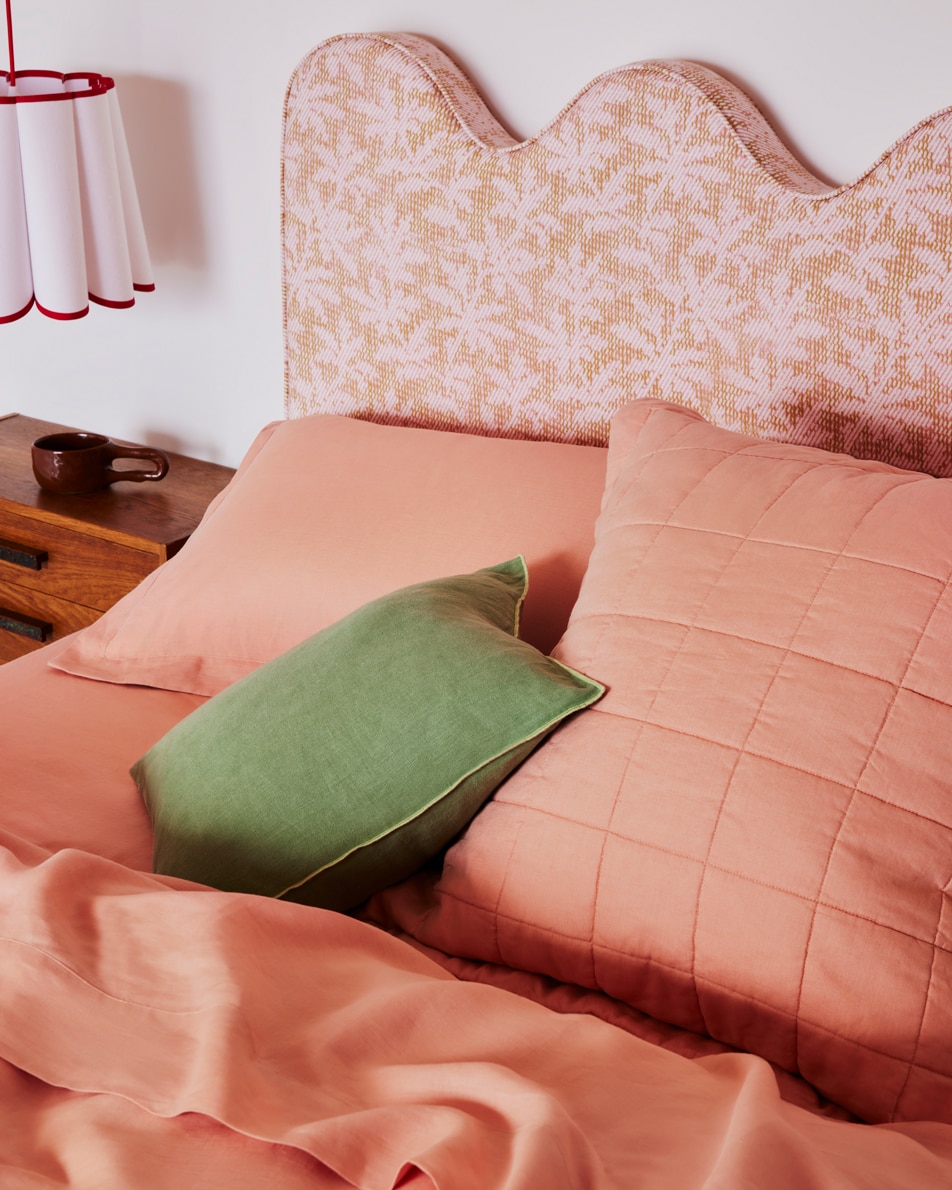 Close up of a bed with pink sheets and a green cushion in front of a pink fabric headboard