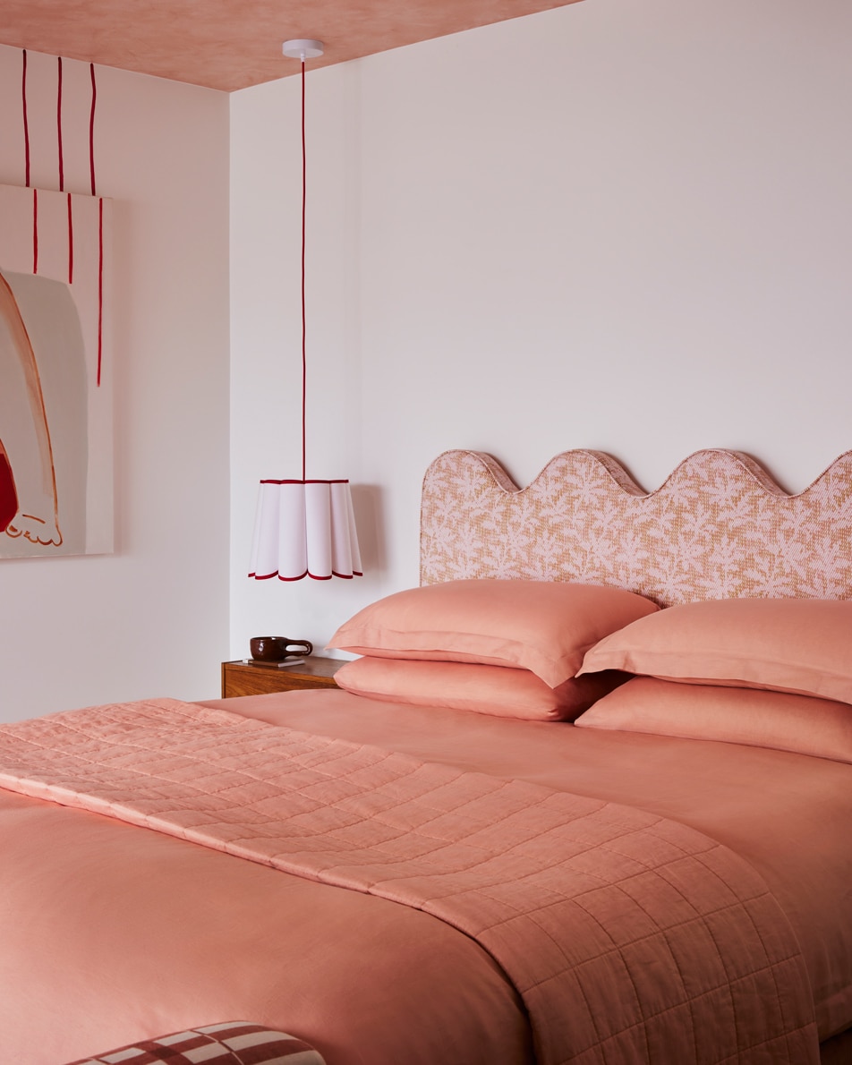 A bed with pink sheets next to a hanging lamp, in a white room with a pink ceiling