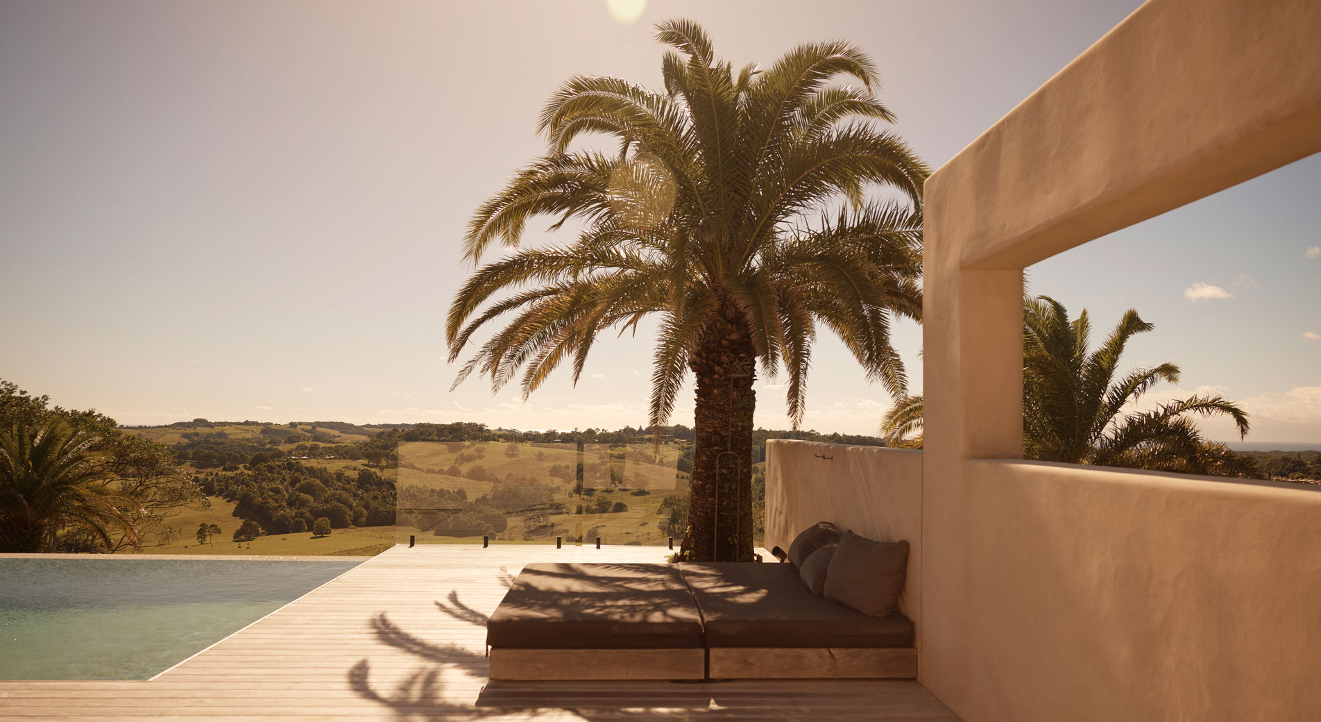 A balcony looking out to grassy hills. In the foreground is a pool and a daybed underneath a palm tree