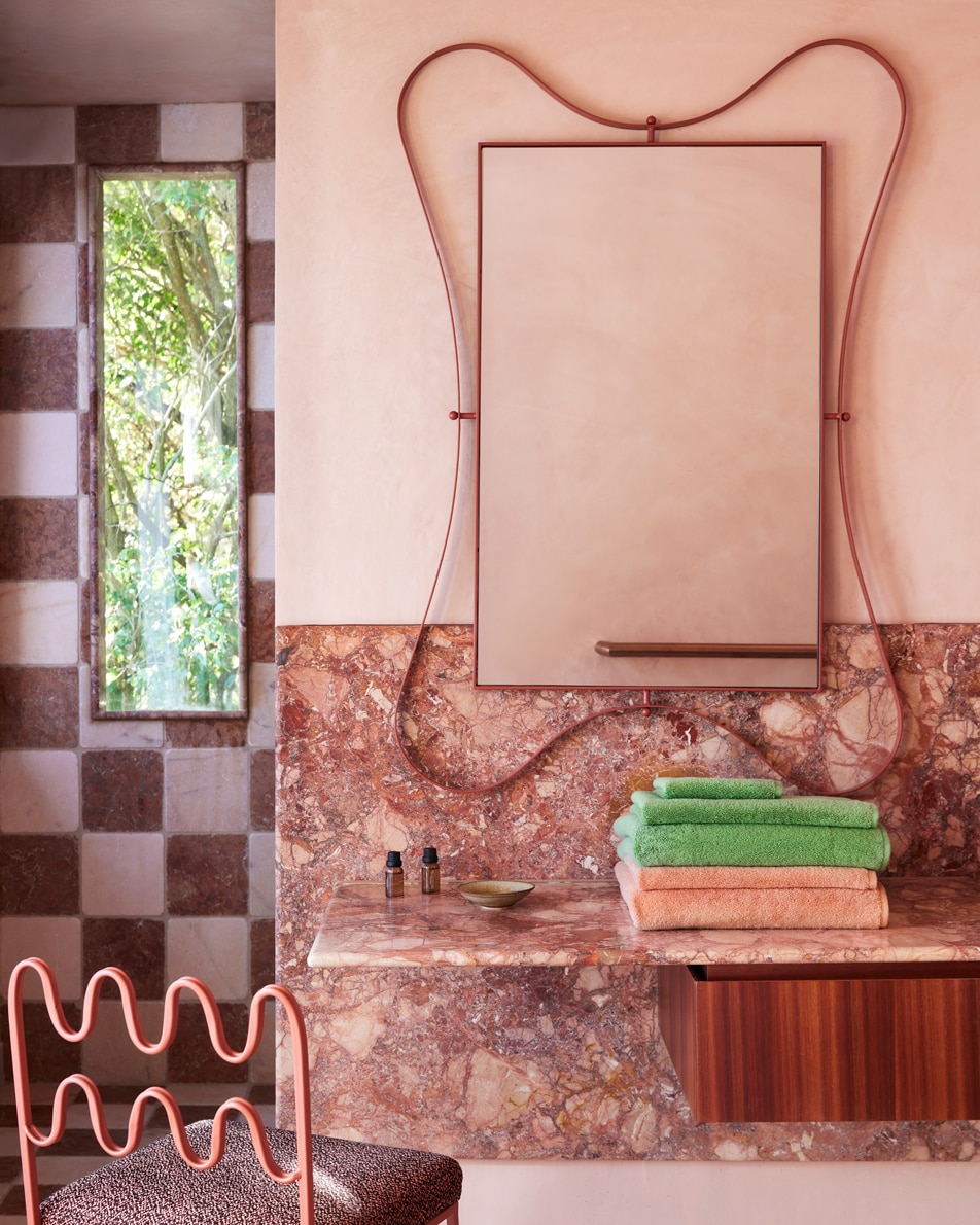 A funky pink bathroom with a wavy mirror on the wall, a stack of towels folded on the pink marble bench and a chequered tiled wall in the background