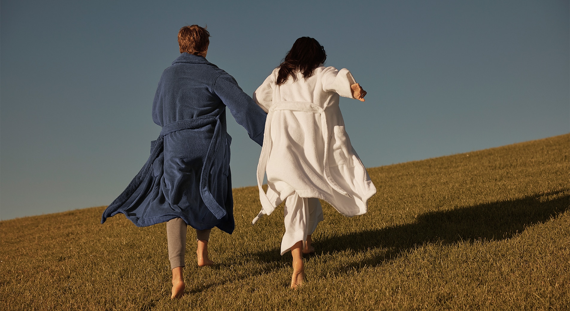 Two people holding hands and running across a grassy hill, both wearing Australian cotton terry bathrobes