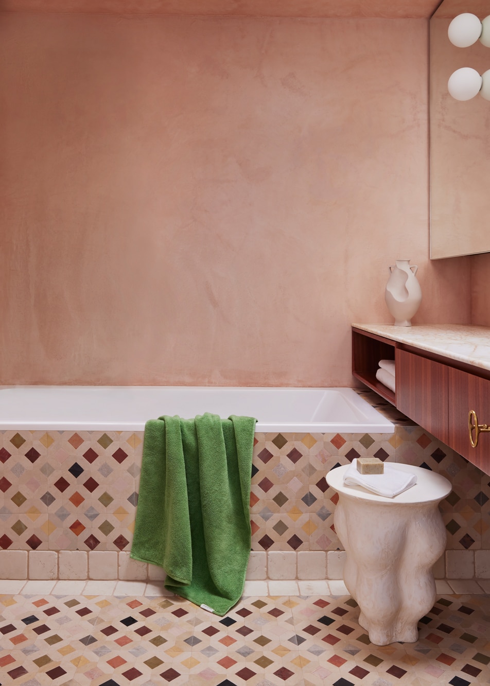 A pink bathroom with colourful diamond floor tiles. A green towel is draped over the side of the bathtub.