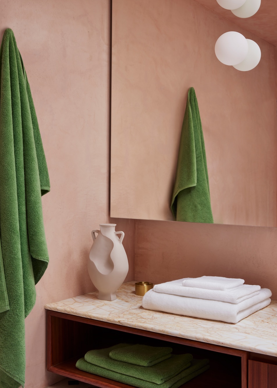 A pink bathroom with a large mirror and marble countertop. A green towel hangs on the wall and a pile of white towels sit on the counter.