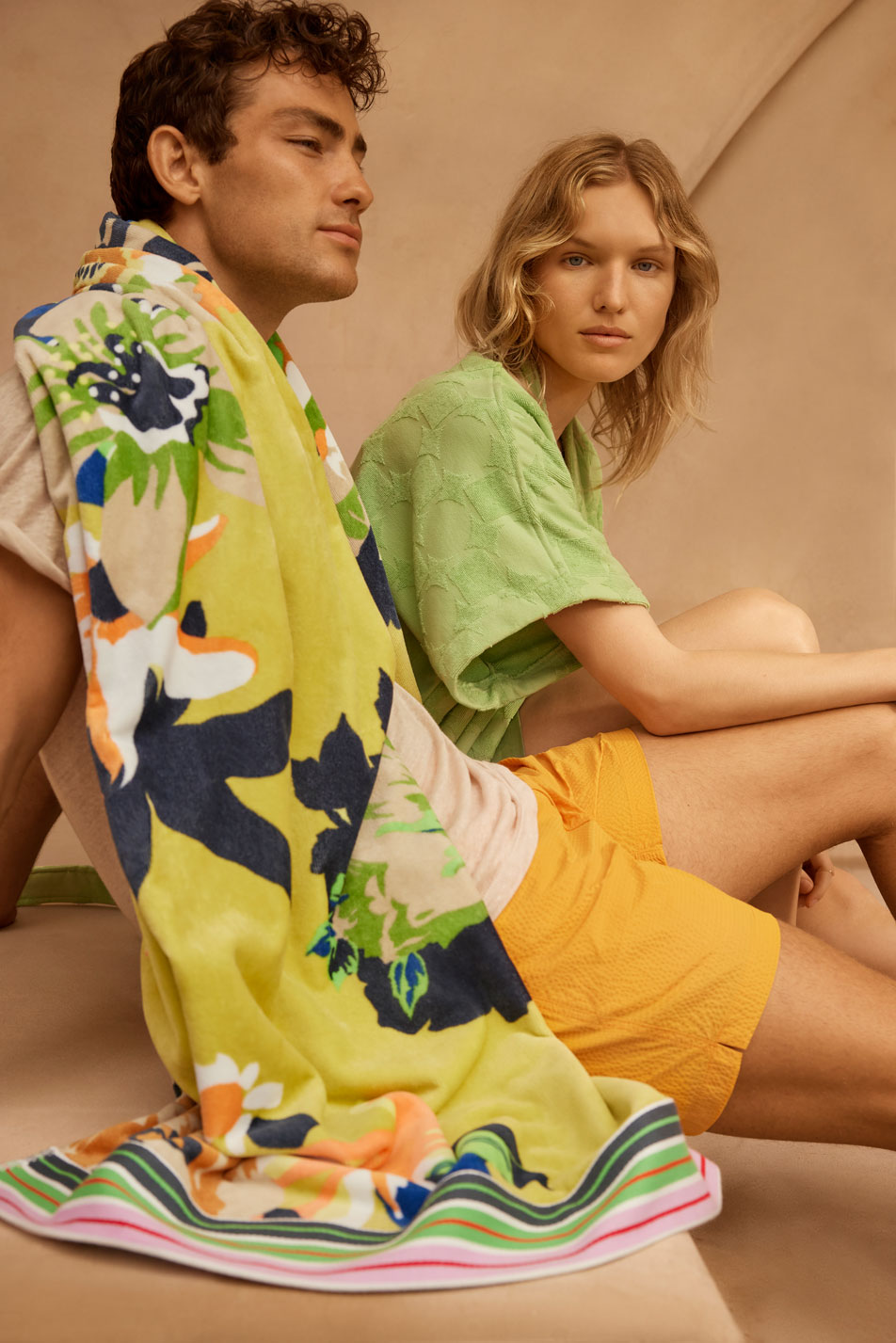 A young brunette man and blonde woman sit side-by-side wearing summer clothing. The man has a bright patterned beach towel draped over his shoulder.