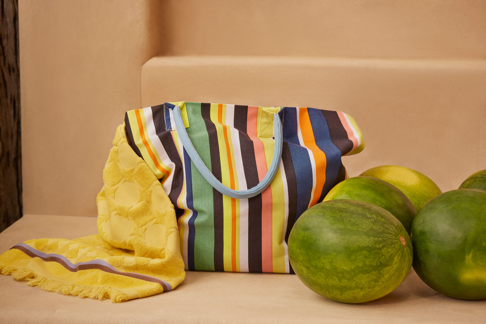 A striped beach bag sitting on a ledge with a yellow beach towel draped over it. Five watermelons sit beside it.