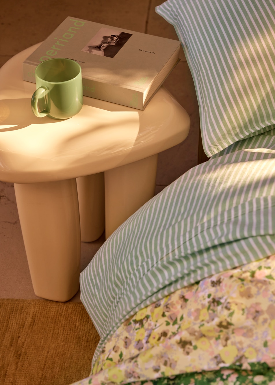 A table next to a bed, on top sits a green glass mug and a book