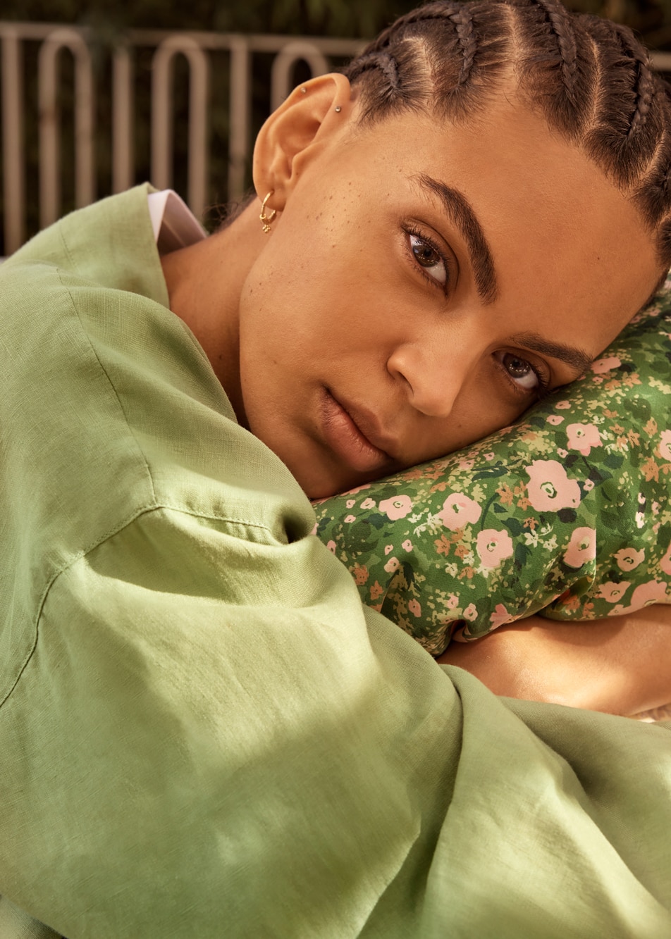 A young person with braids wearing a green linen robe, resting their face on a floral print quilt