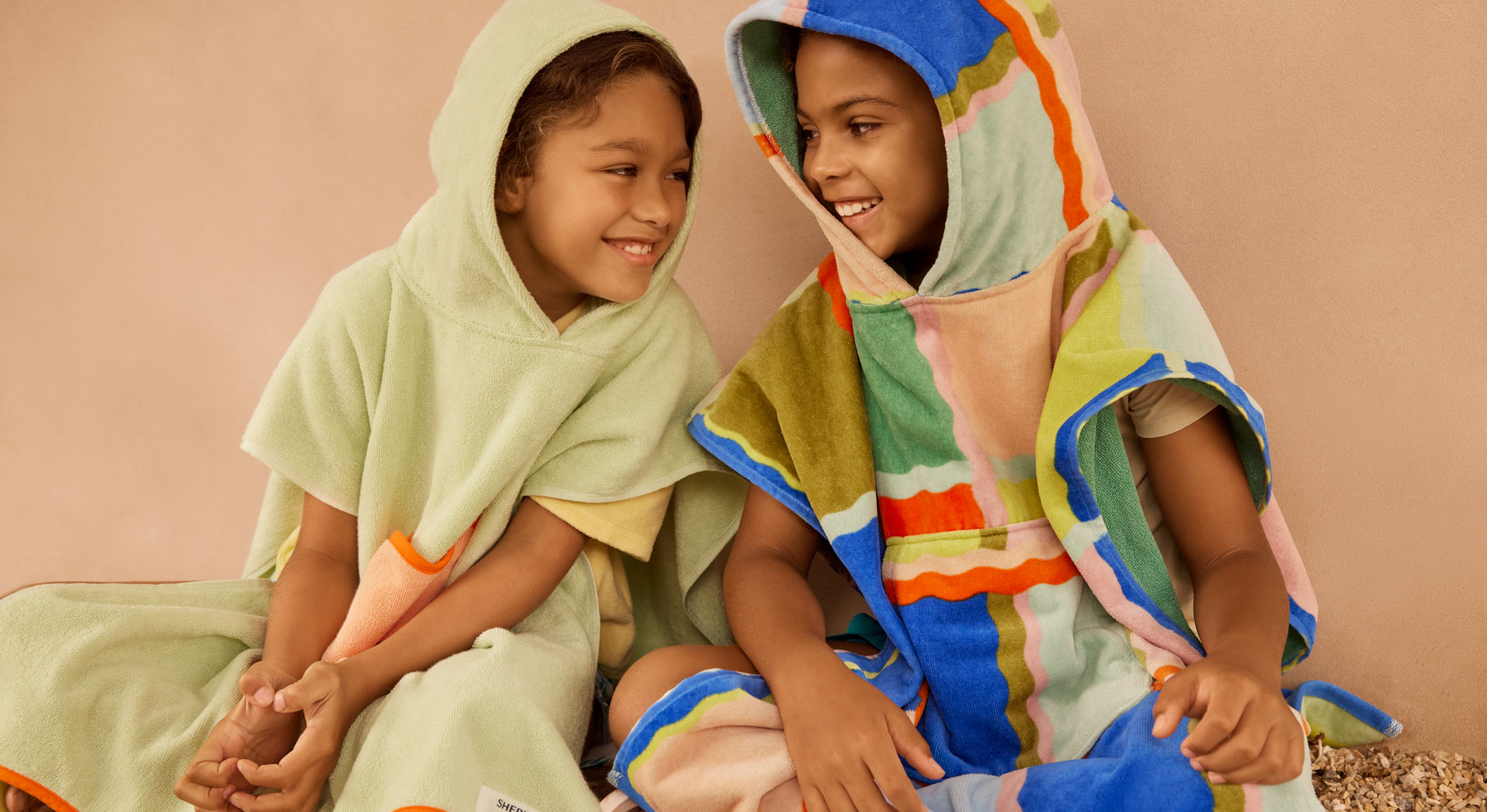 Two young kids wearing Sheridan beach ponchos, sitting beside each other and smiling