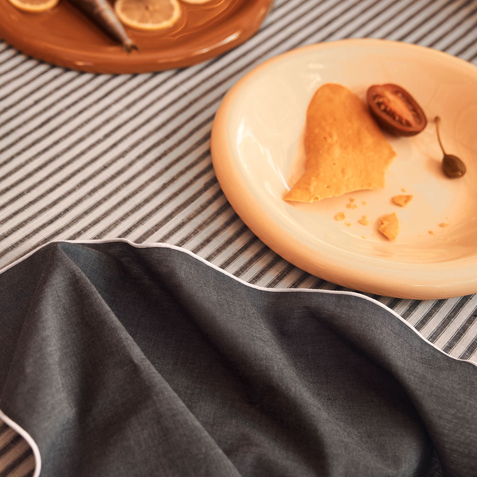 square image, close up of a napkin laying tousled on top of a ticking stripe tablecloth. Two ceramic plates of food sit on the table.