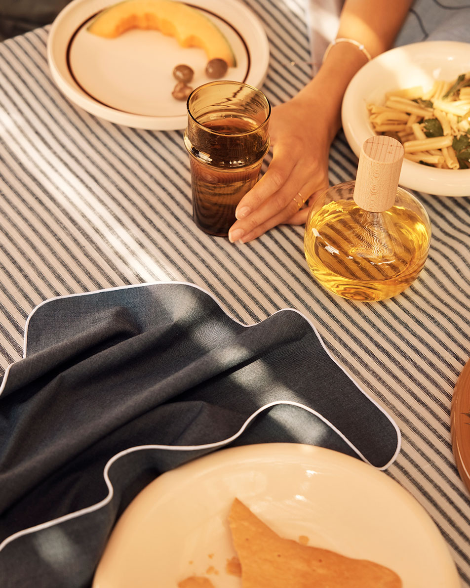 Close-up photo of a dining table, dressed in a ticking stripe tablecloth with a carbon linen napkin on top. Plates of food and a bottle of olive oil sit on top, next to a hand reaching out for a glass of water.