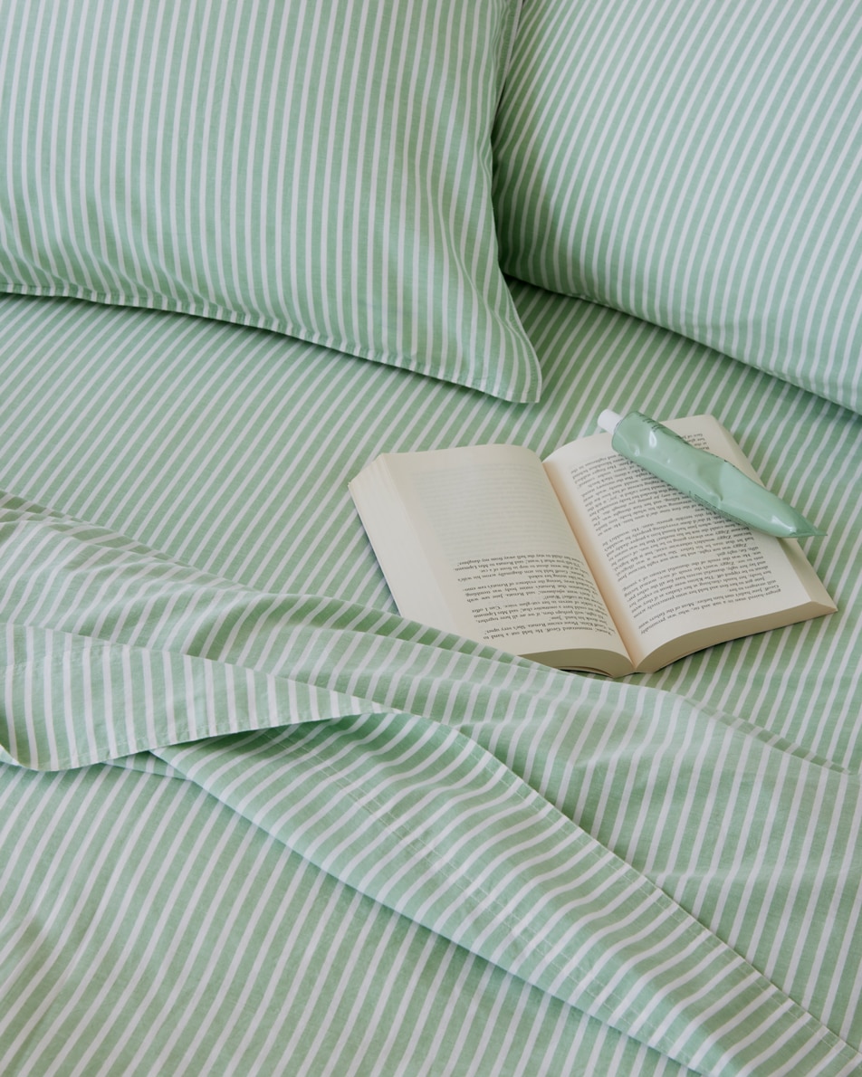 A close-up of a green striped bed. An open book and a tube of hand lotion sit on top of the tousled sheets.