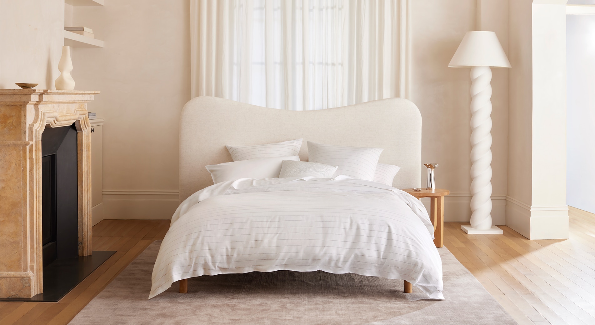 A bedroom with white walls, sheer fabric curtains and timber floors. An all-white bed sits in the middle on top of a grey rug. To the left is a marble fireplace and to the right is a tall lamp.