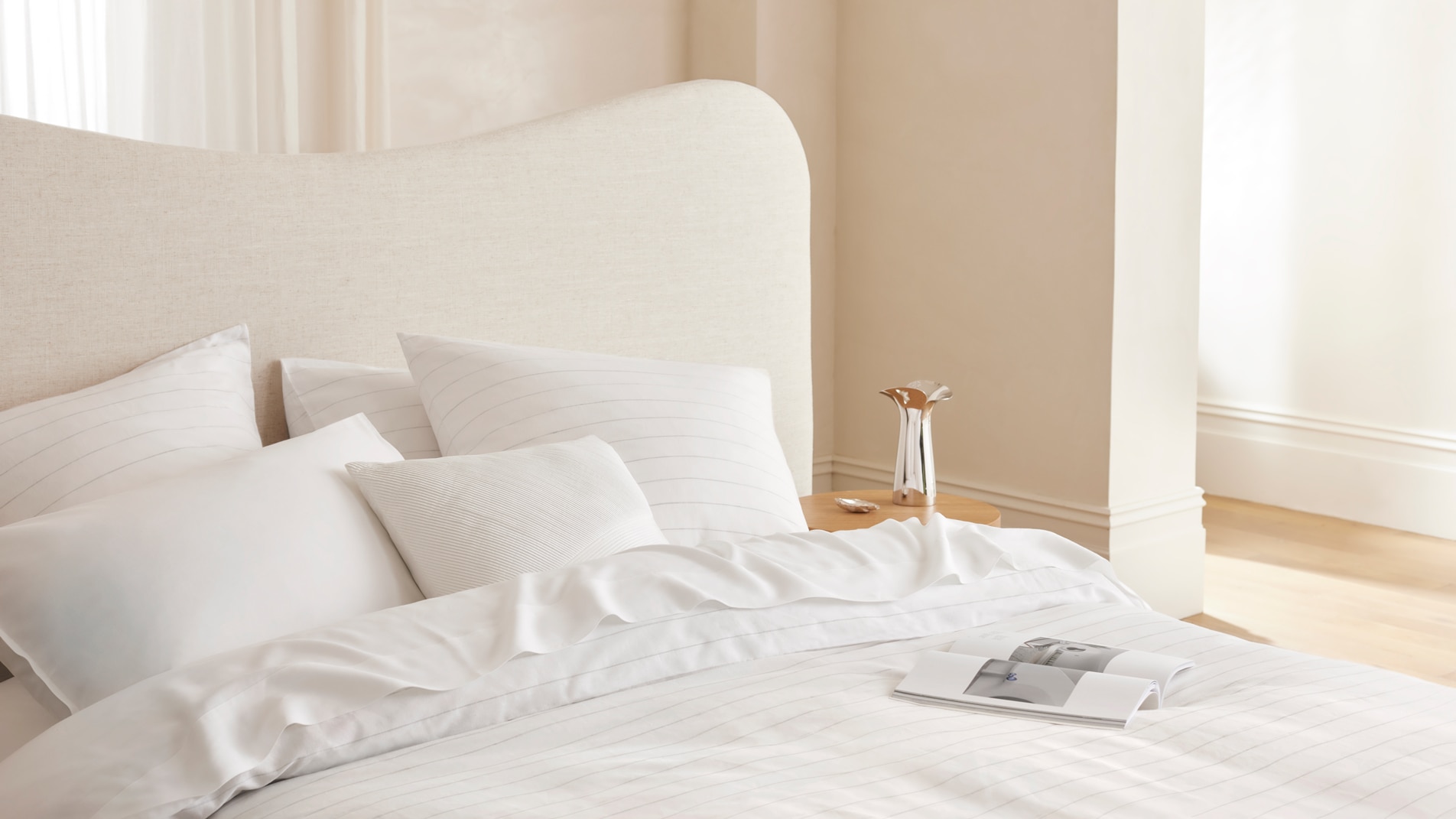 A close up of a bed with all white bed linen and a magazine sitting on top. In the background a silver vase sits on a round timber table.