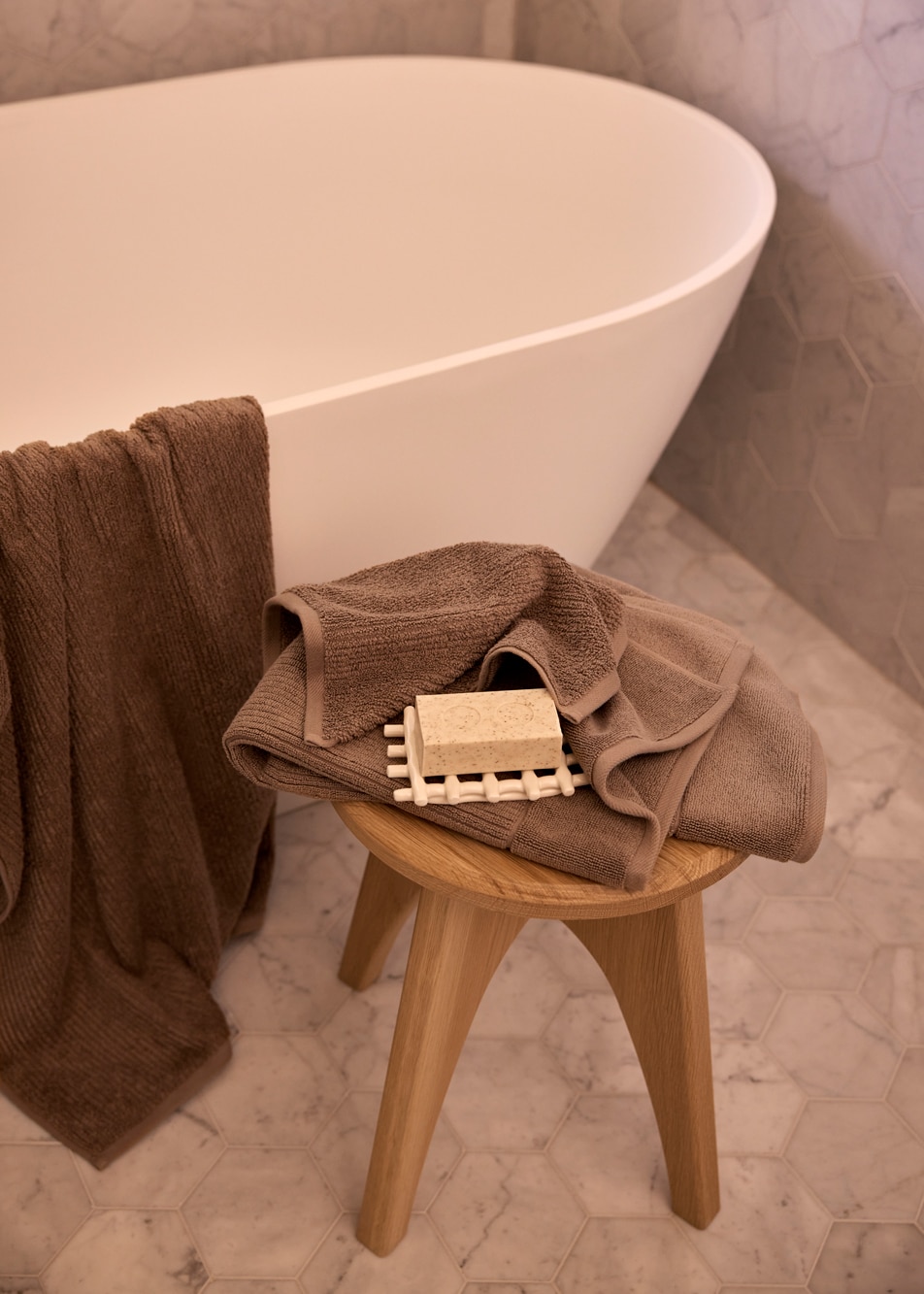 A marble bathroom. There is a bathtub with a towel draped over the side, and a timber stool with a towel and bar of soap sitting on top.