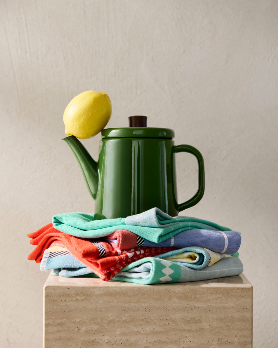 A green kettle sits on top of a stack of Sheridan tea towels, with a lemon balancing on its spout