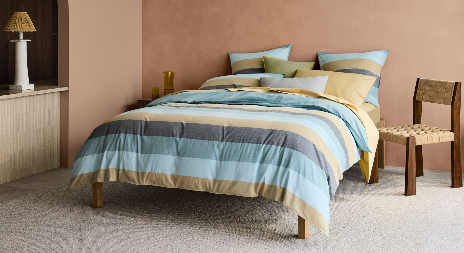 A blue and ochre striped bed sitting in a bedroom with peach coloured walls. Next to the bed is a wicker chair and a white lamp.