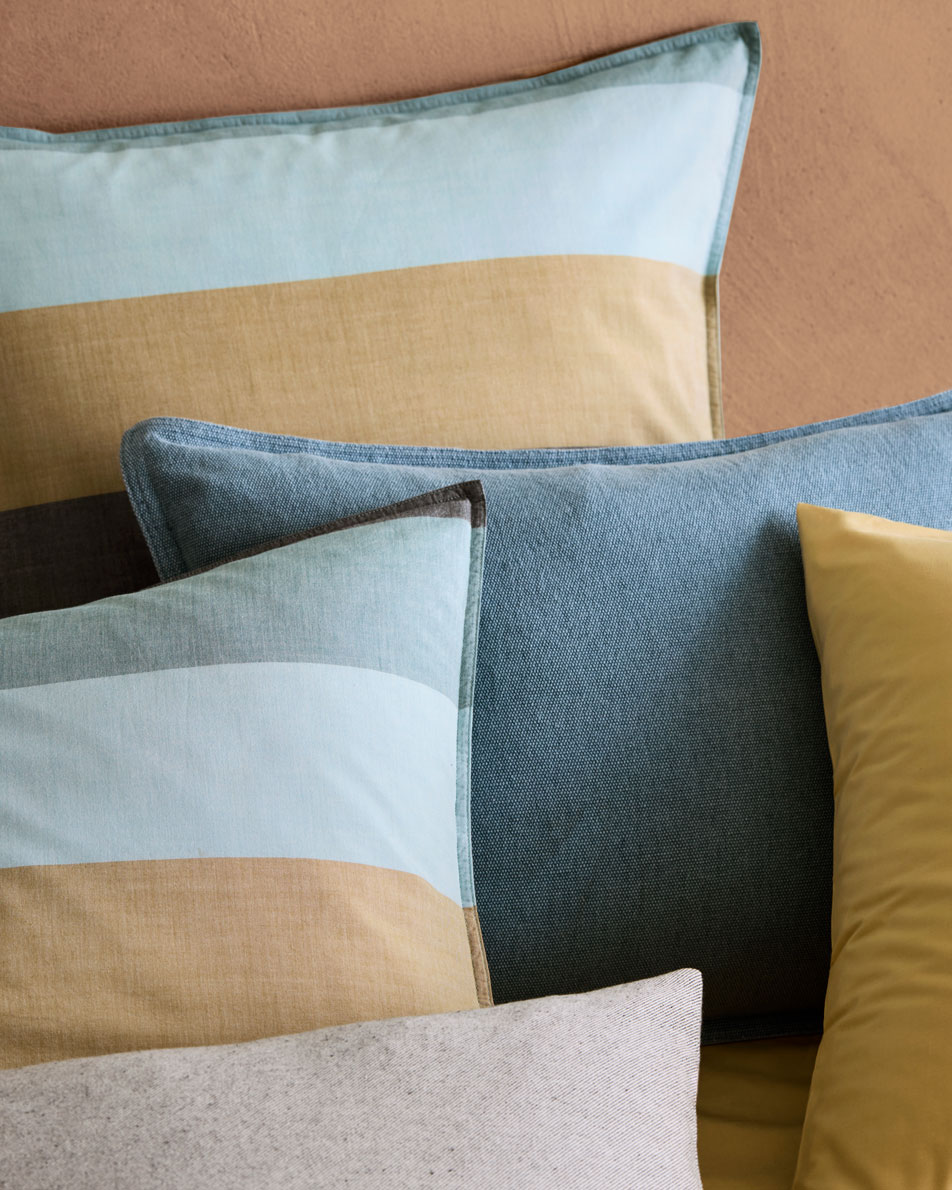 A close-up of layered pillows. Some have blue and ochre striped pillowcases and some are plain dye in coordinating colours.