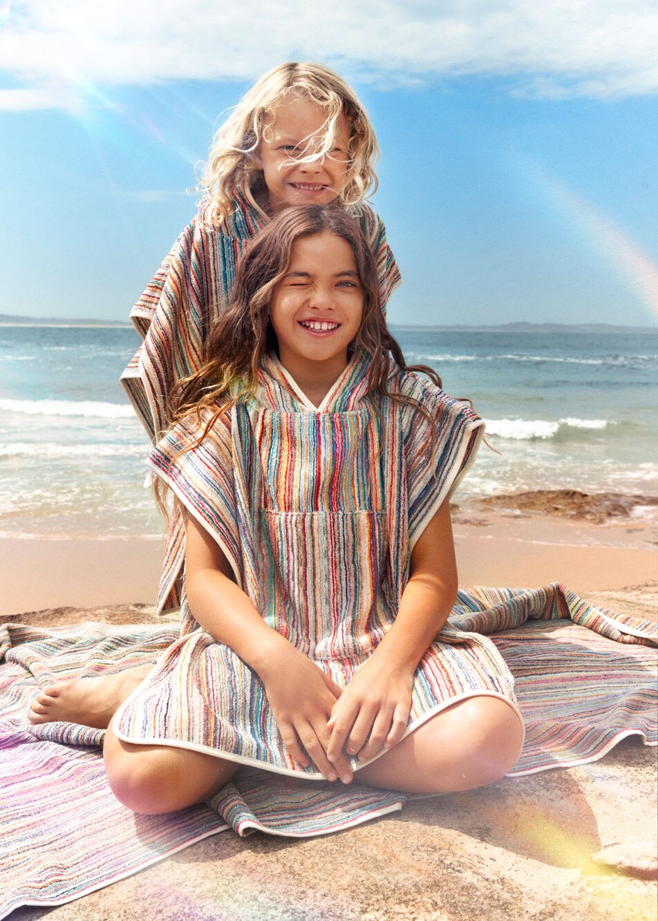 Two children smiling on the sand at the beach. Both are wearing Sheridan Cancun beach ponchos and are sitting on a Cancun beach towel.