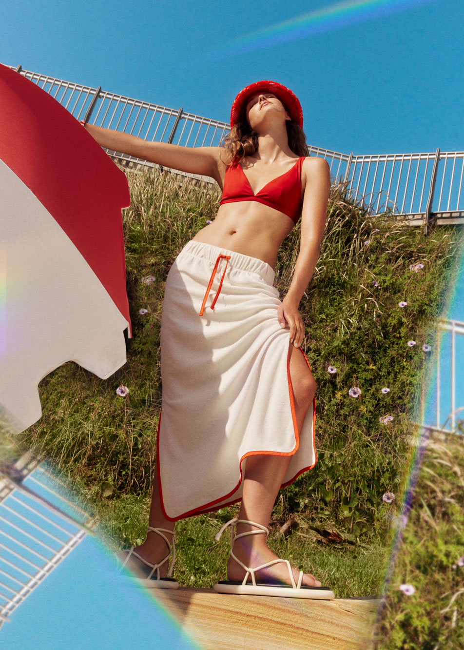 A young woman wearing Sheridan beach clothing, standing outside by flowering plants and a beach umbrella