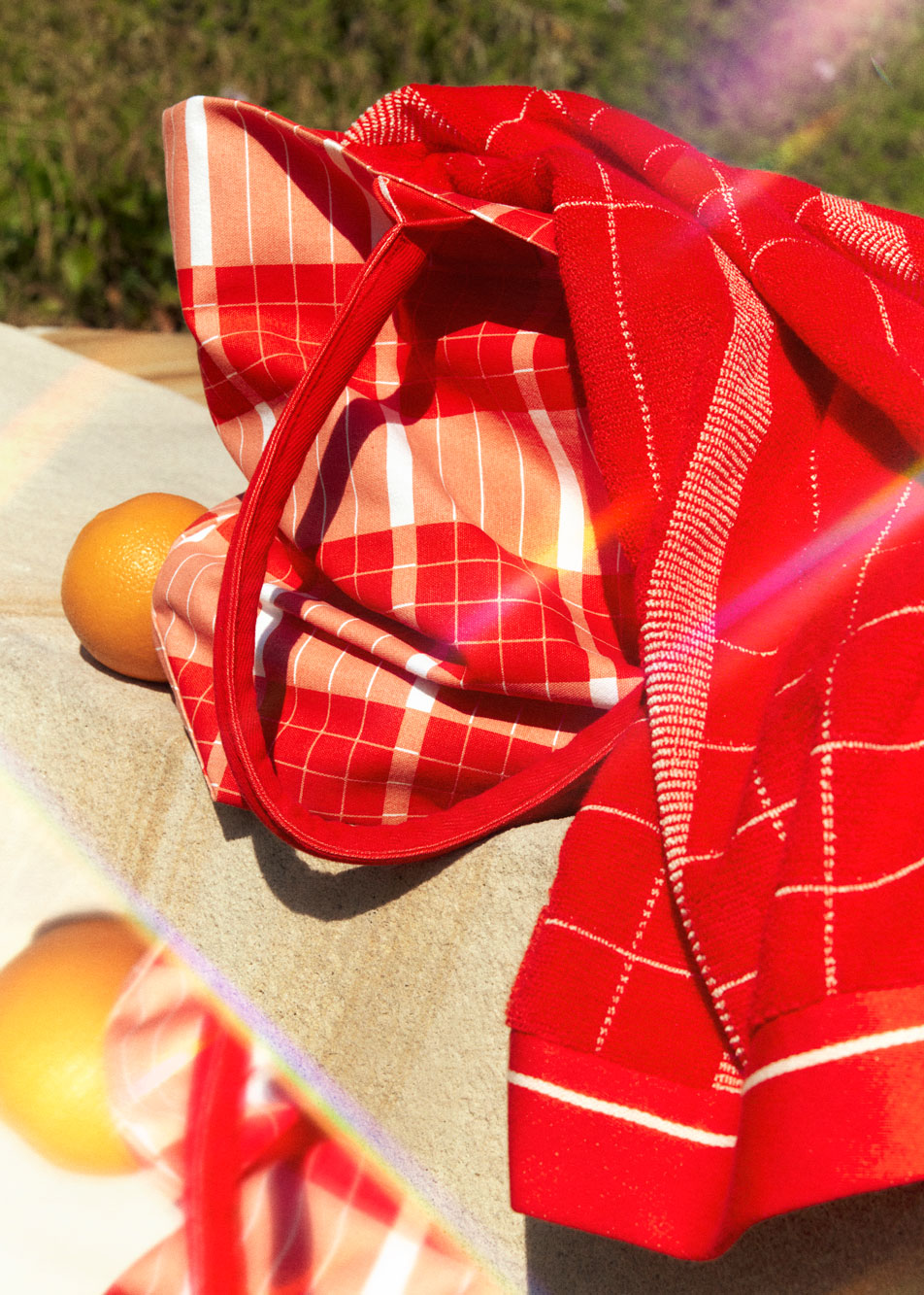 A Sheridan beach bag sitting on an outdoor fence. A Sheridan beach towel spills out of the top of the bag, with oranges sitting next to it.