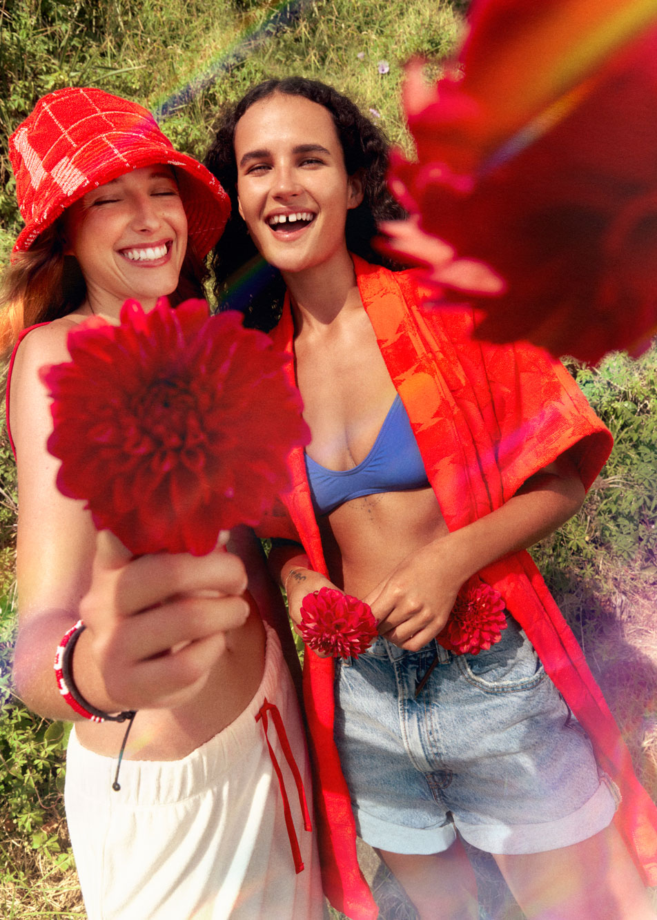 Two young women smiling at the camera, standing side by side. The woman on the left is holding a red flower out towards the camera.