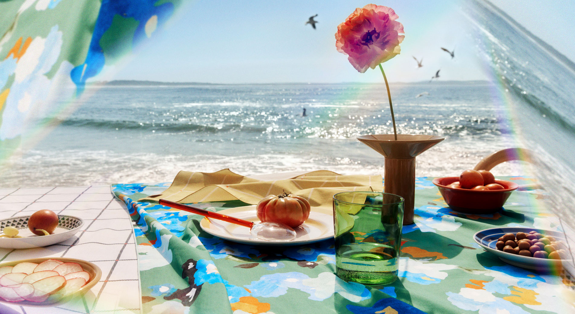 photo of styled table by the beach. A green floral tablecloth with plates of vegetables on top, and a single pink flower in a ceramic vase.