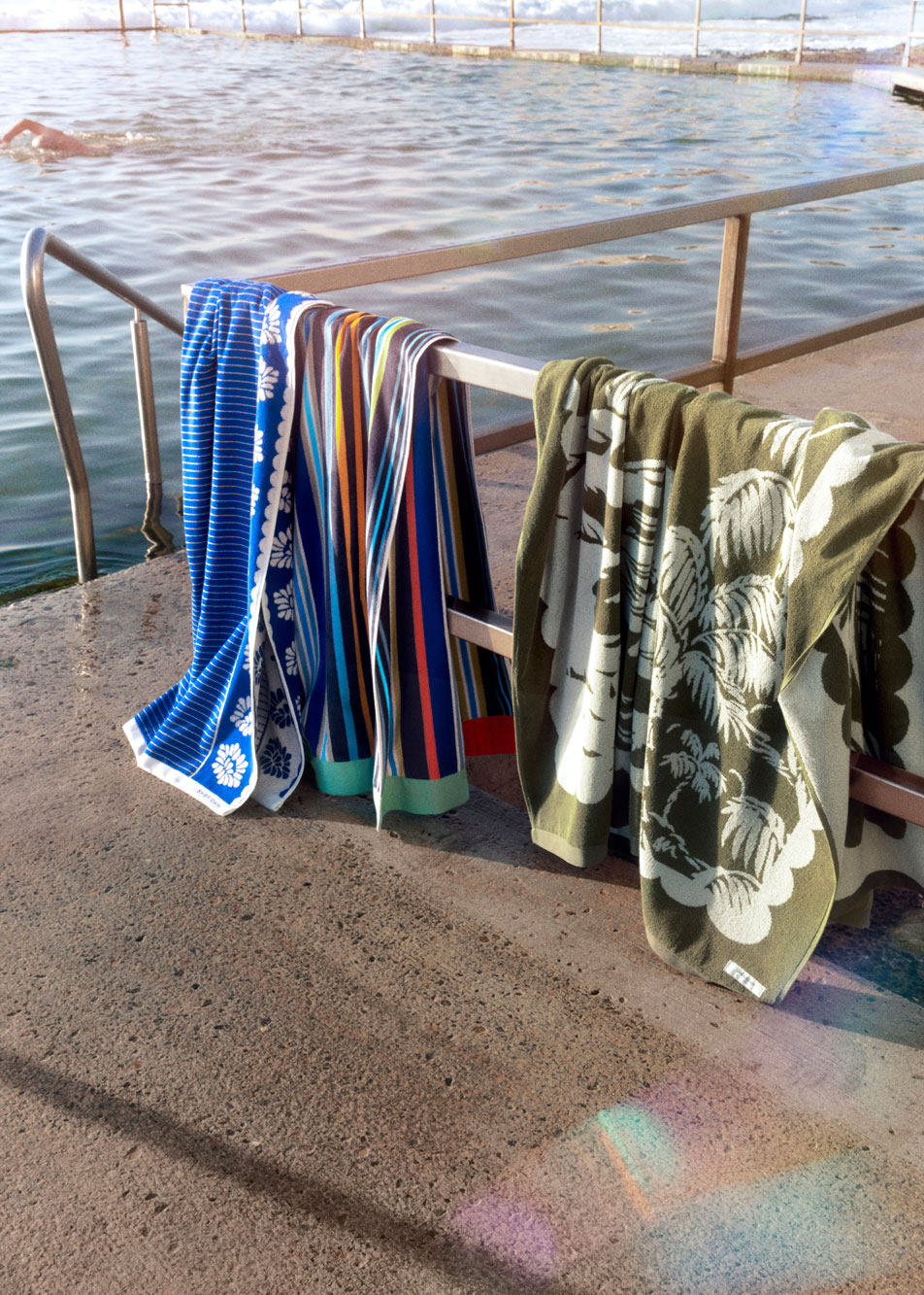 Four Sheridan beach towels in different designs hanging on a fence by the beach.