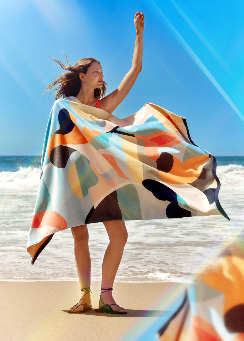 A young woman standing in front of the ocean at the beach, holding a Sheridan beach towel that's blowing in the wind.