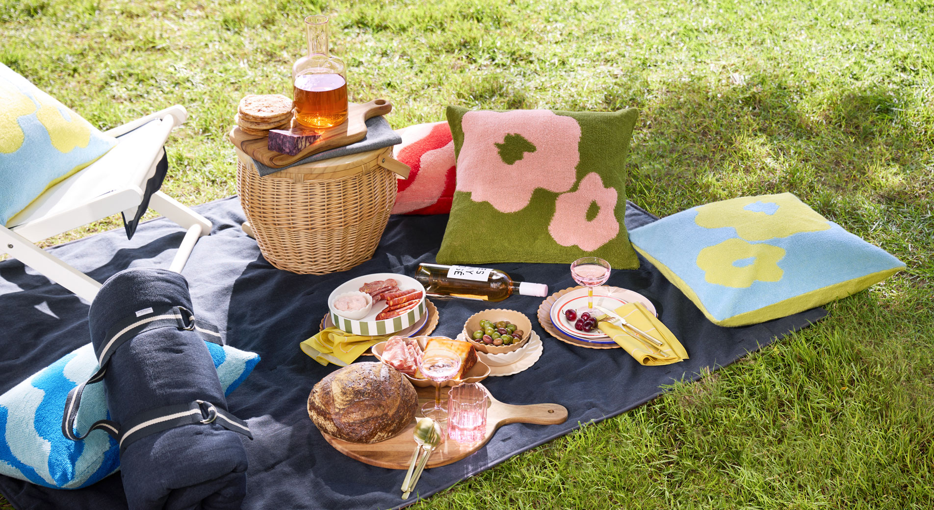A picnic set up on the grass, with cushions, a picnic rug and various foods and drink