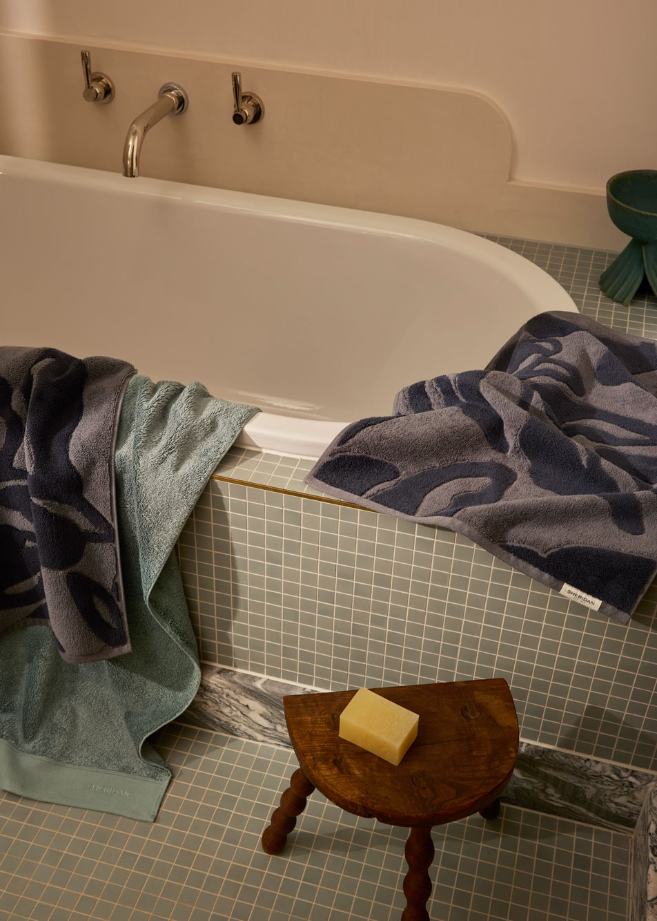 A green tiled bath tub with sea green and charcoal coloured bath towels draped over the side