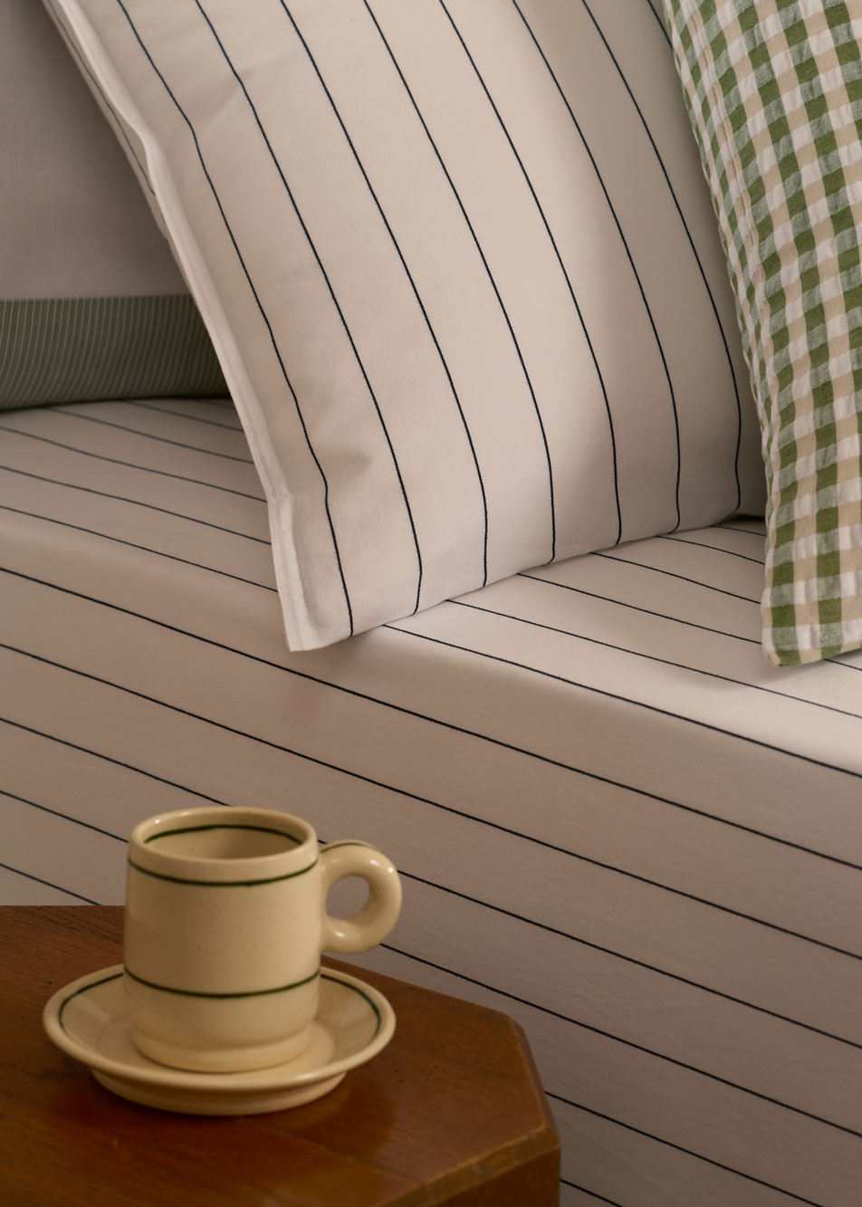 A close-up of a bed styled with Bayley Stripe Washed Percale sheets in white. Next to the bed is a wooden side table with a ceramic mug and saucer.