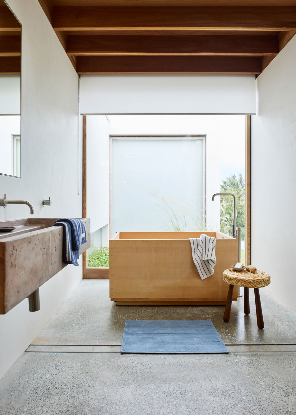 A modern bathroom with a concrete wall-mounted basin, timber bath tub and floor-to-ceiling window at the rear. 