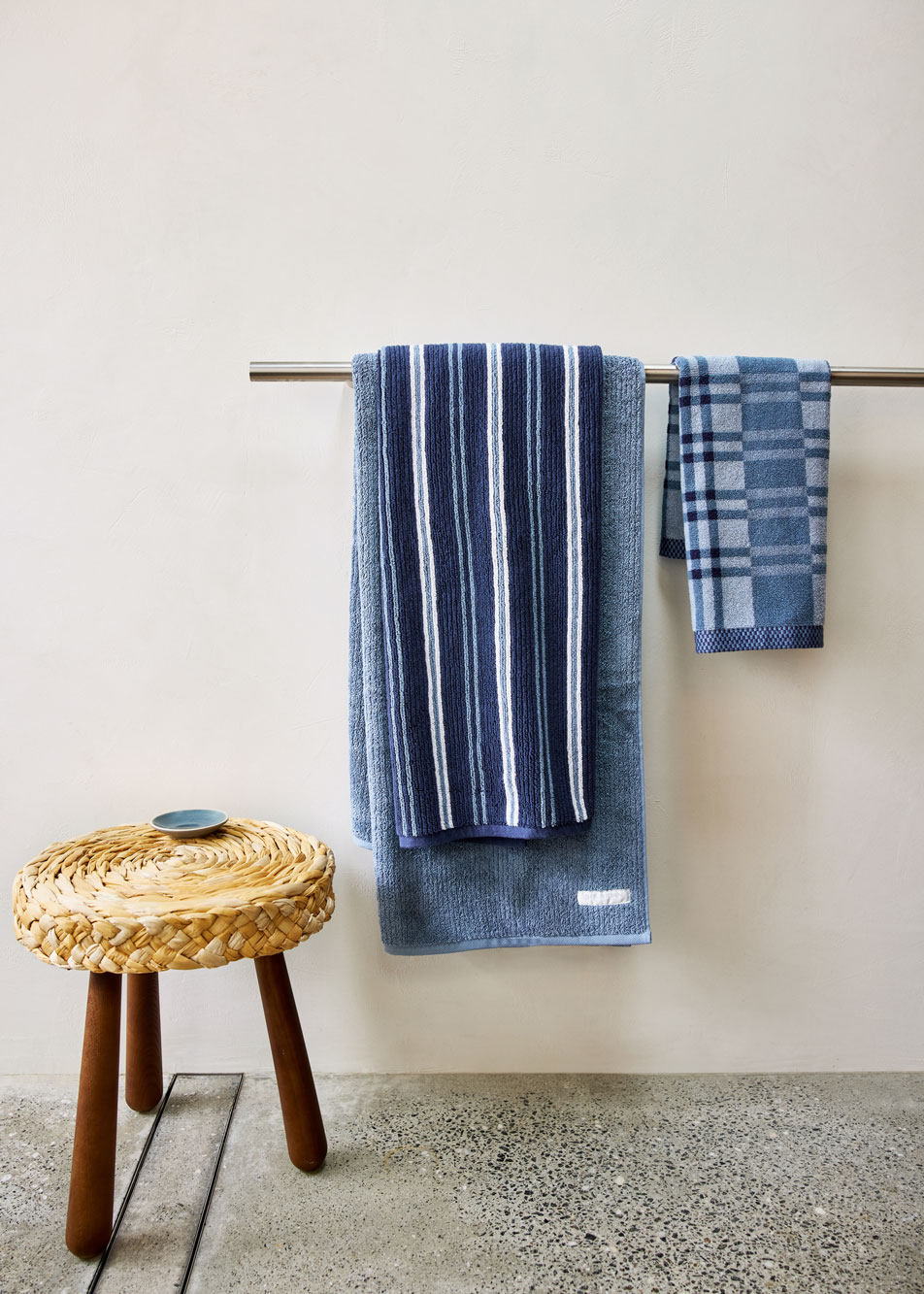 Three blue towels in different stripe and check patterns, hanging on a towel rail. Next to the towels is a wicker stool.