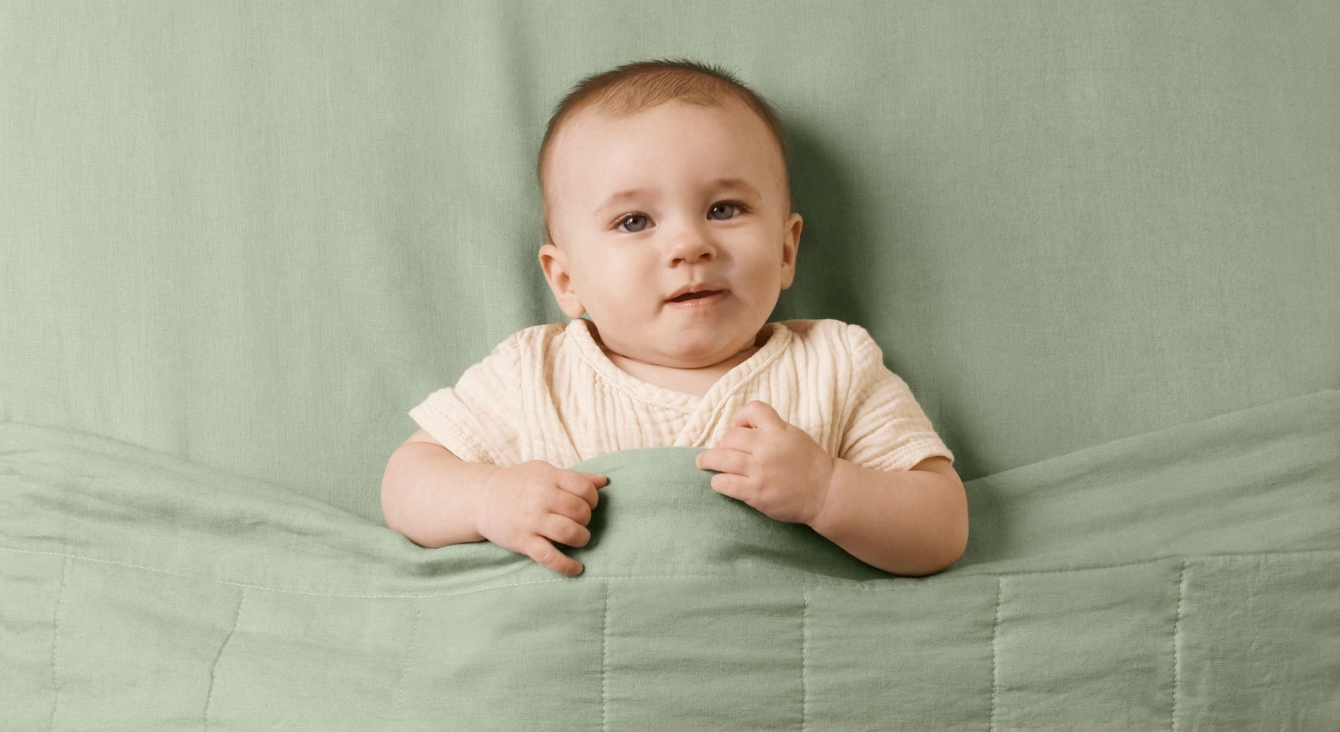 baby in neutral clothing lies in cot, with green abbotson fitted cot blanket and linen baby blanket. arms are over blankets edge.