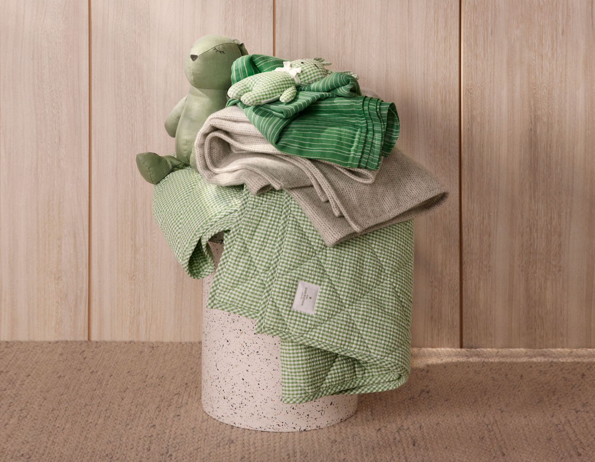 marble plinth on carpeted floor, with wood background. stacked on top is green checked baby blanket, grey wool knit blanket, striped green blanket, and two baby toys.