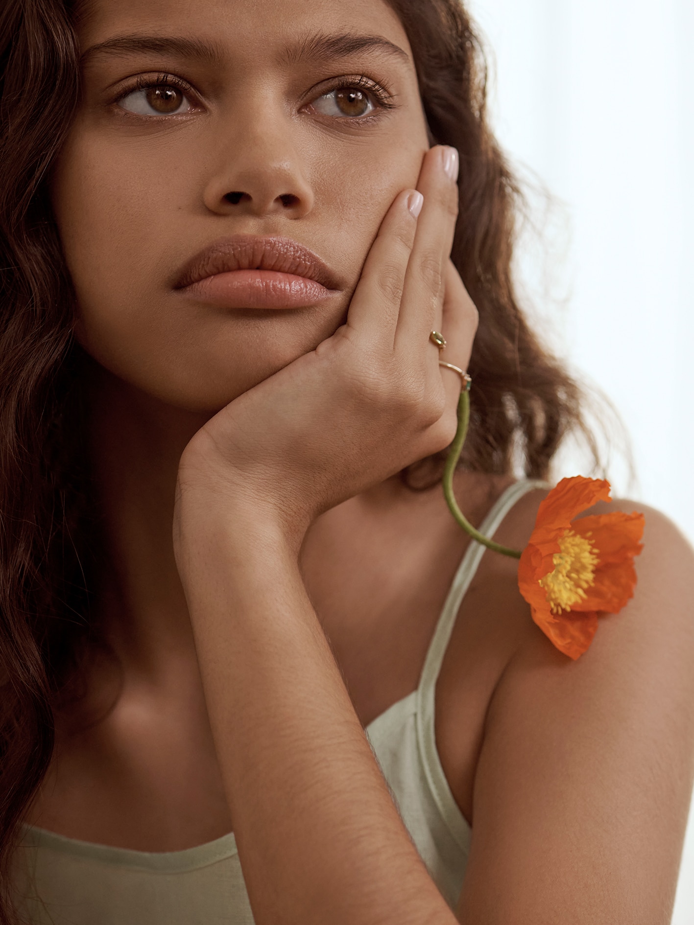 brown woman sits with her chin in her hand, with a flower held between two fingers. she wears a julip linen slip dress.