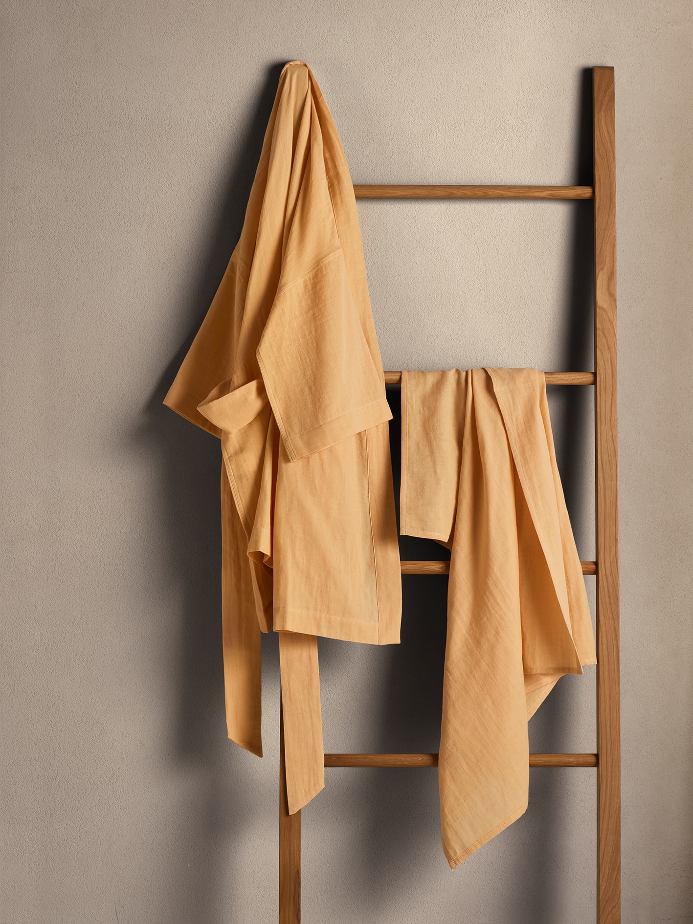 biege colourd wall, with brown wooden ladder hanging from it. rockmelon coloured belgian flax linen robe and wrap are draped on top of ladder and rung, respectively. r