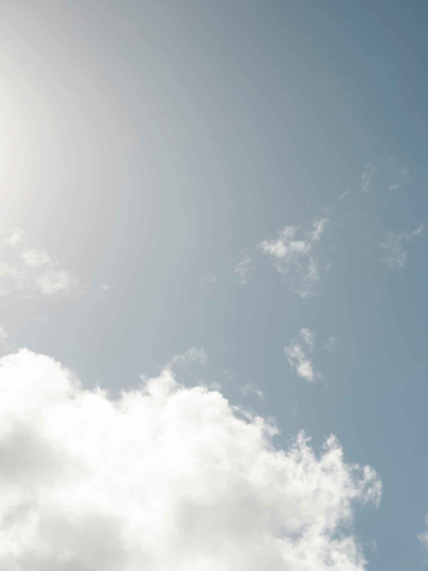 shot of blue sky with white puffy cloud coming through from bottom left hand corner of photo. light from sun is seen, but not sun itself.