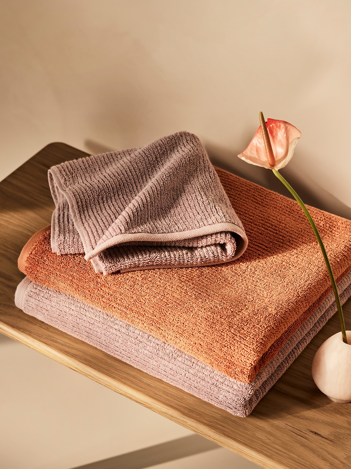 Brown and orange stacked cotton bath towels on wooden beach, with single flower in vase sitting next to it.
