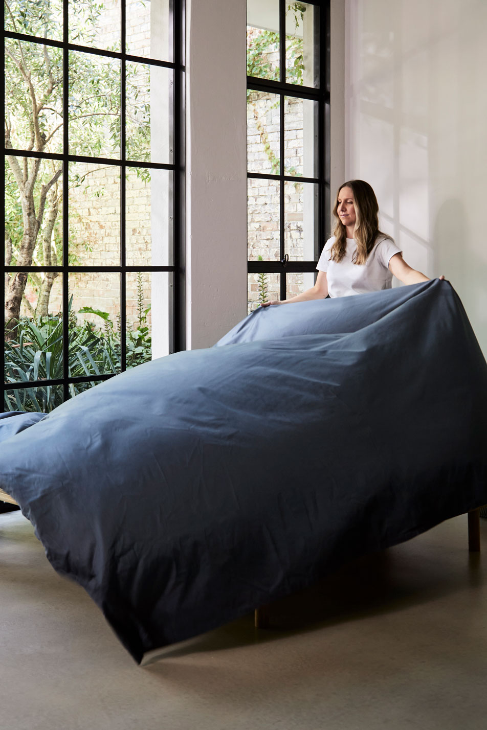 A young woman styling a bed with navy blue cotton sateen sheets. In the background are tall French windows with trees outside.