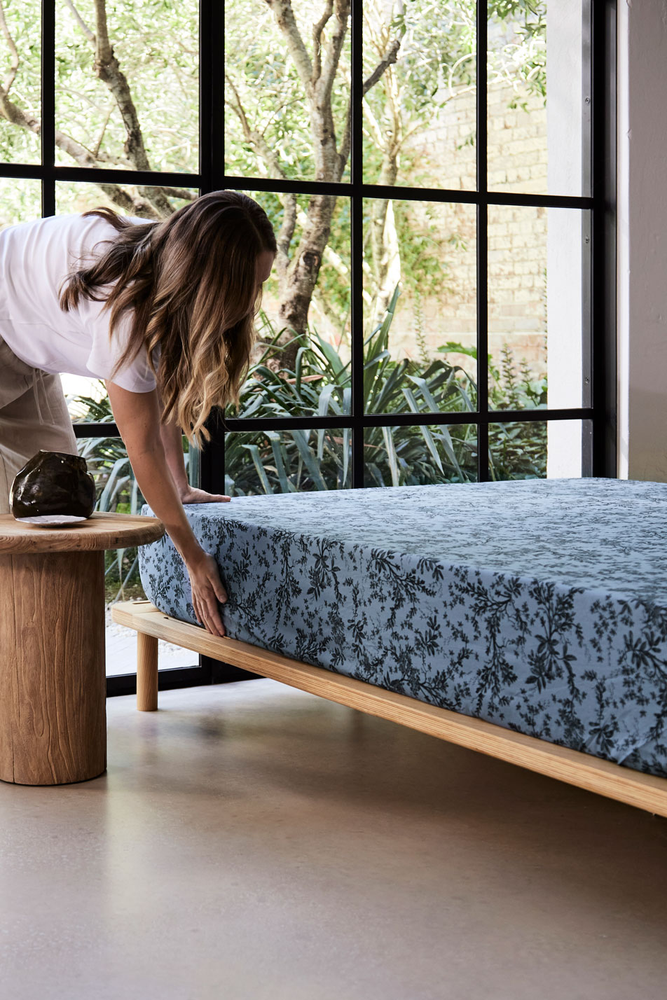 A young woman styling a floral print fitted sheet on a mattress. Next to the bed is a timber side table and in the background are tall French windows with trees outside