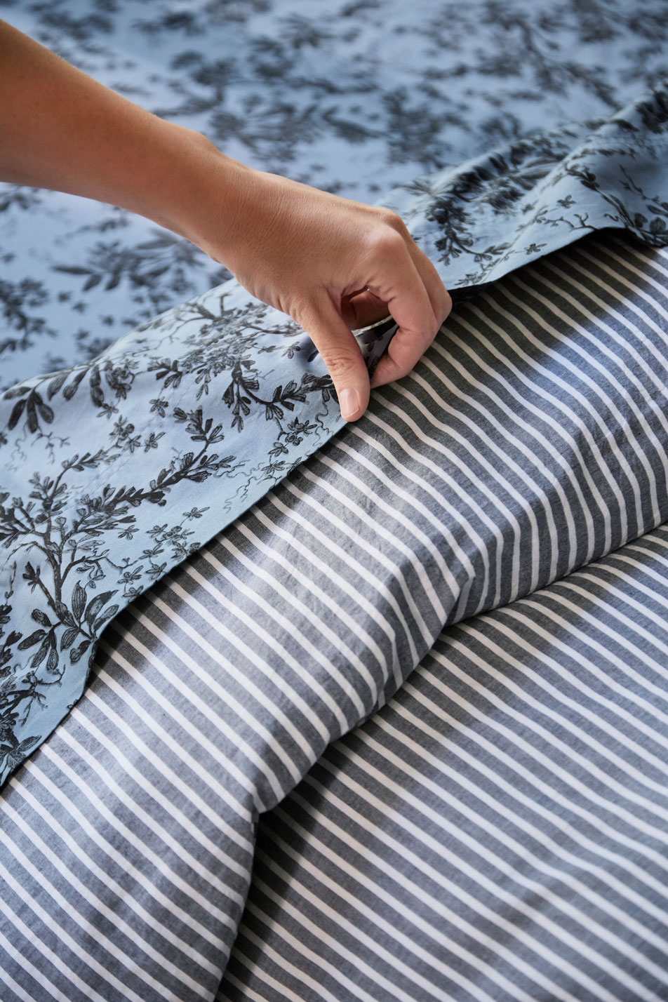 A close-up of a woman's hand making a bed. The bed has floral sheets and a striped quilt cover.