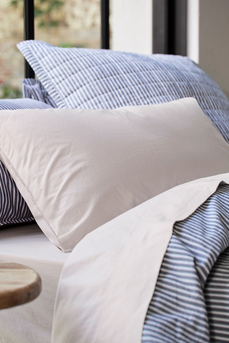 A close-up of pillows on top of a bed, dressed in neutral sheets and a striped quilt